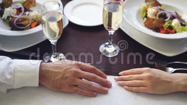 在优雅的餐厅里，年轻情侣触摸和牵手的特写镜头。 提供起泡香槟杯的桌子